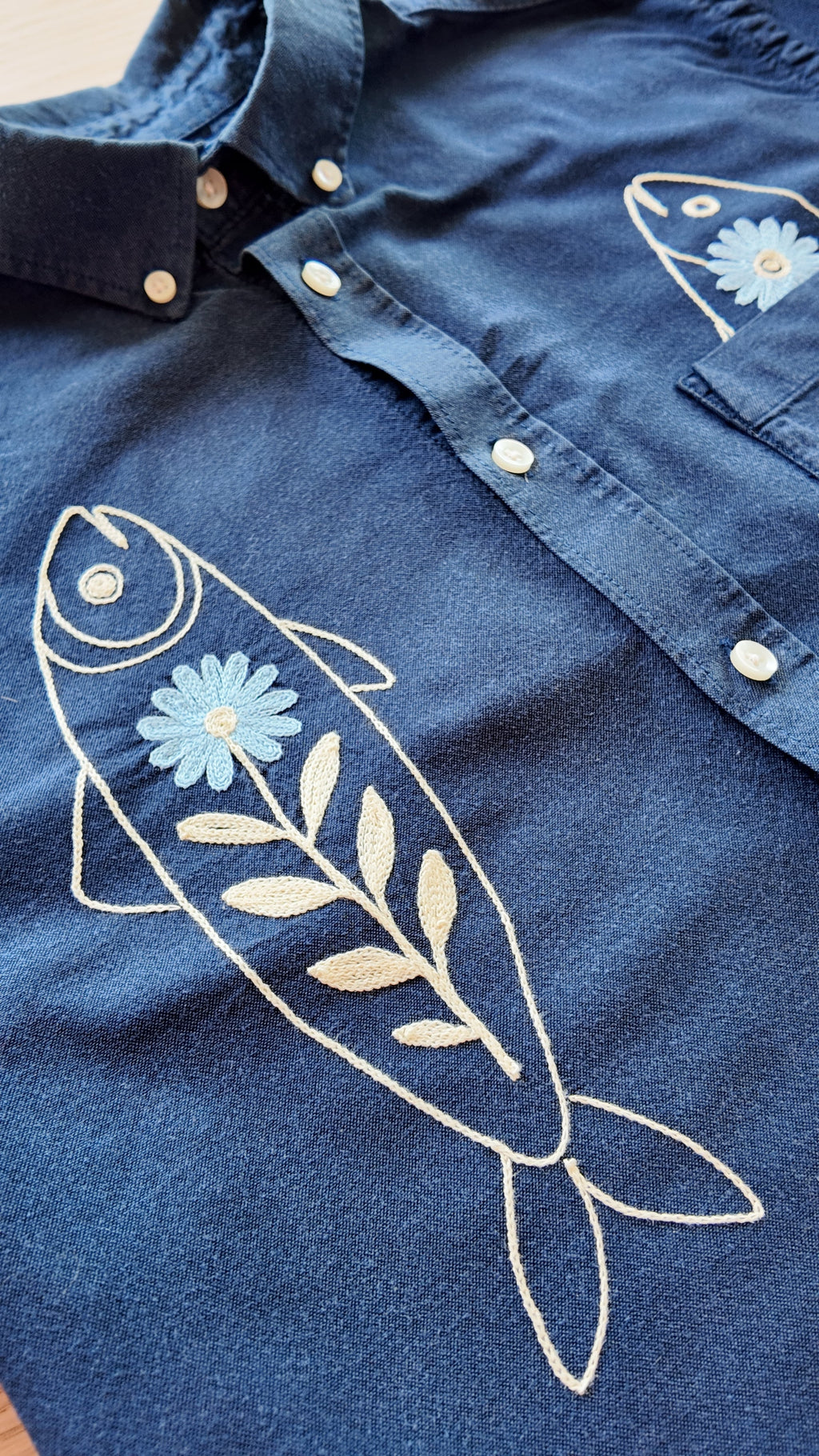 Close-up of off white fish and blue flower chainstitch embroidery detail on navy blue button-down