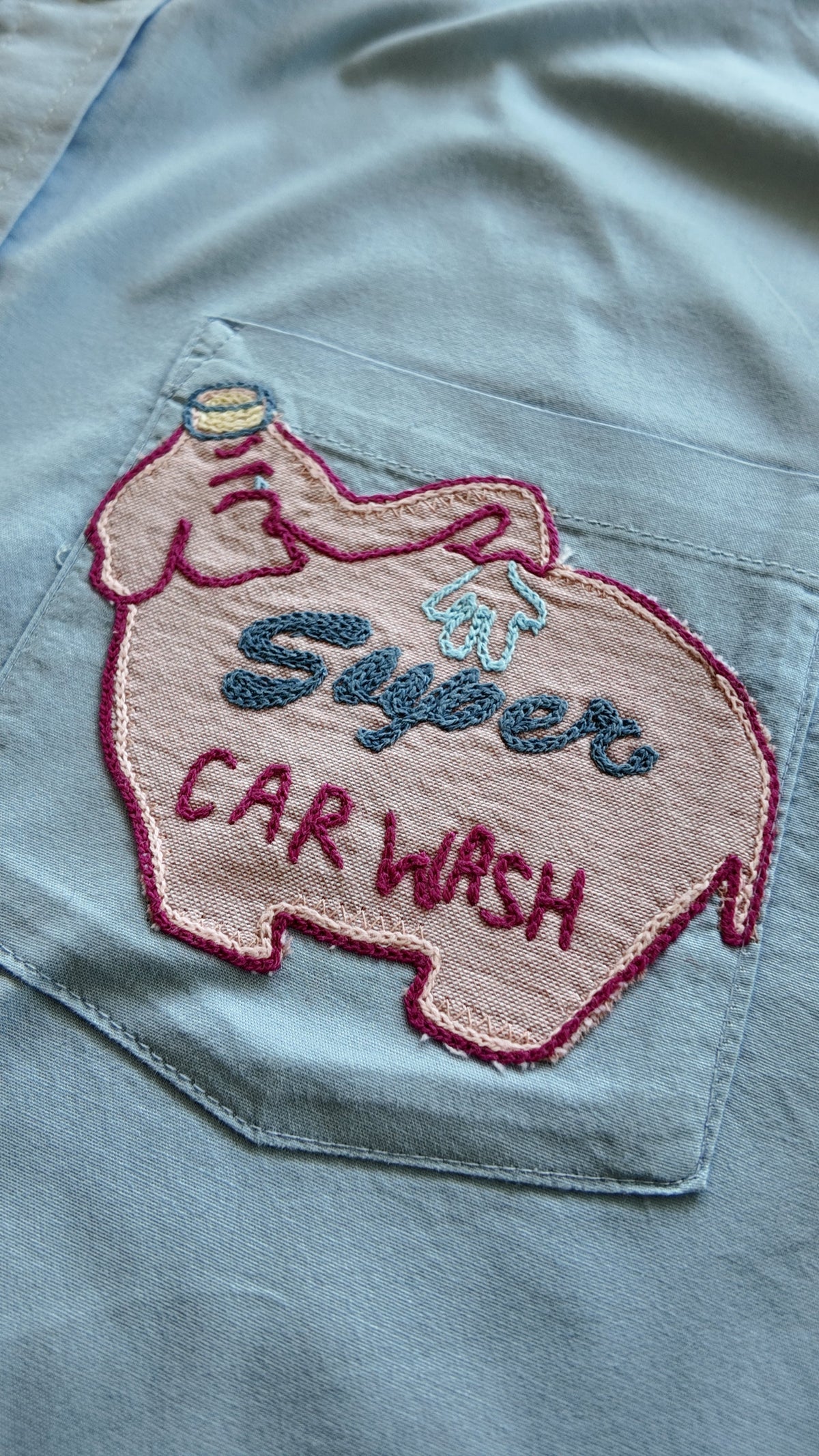 Close-up of pink car wash chainstitch embroidery detail on pale blue button-down pocket