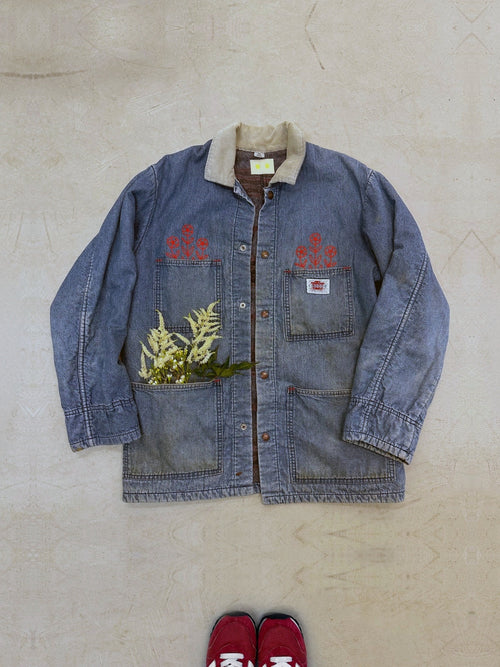 One-of-one vintage Caboose denim chore coat in mid-wash indigo with hand-chainstitched red floral embroidery growing from the front chest pockets